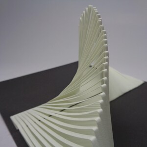 May include: A white, abstract sculpture with a curved, fan-like design. The sculpture is made of layered, thin, white material, creating a sense of depth and texture. It sits on a dark gray surface.