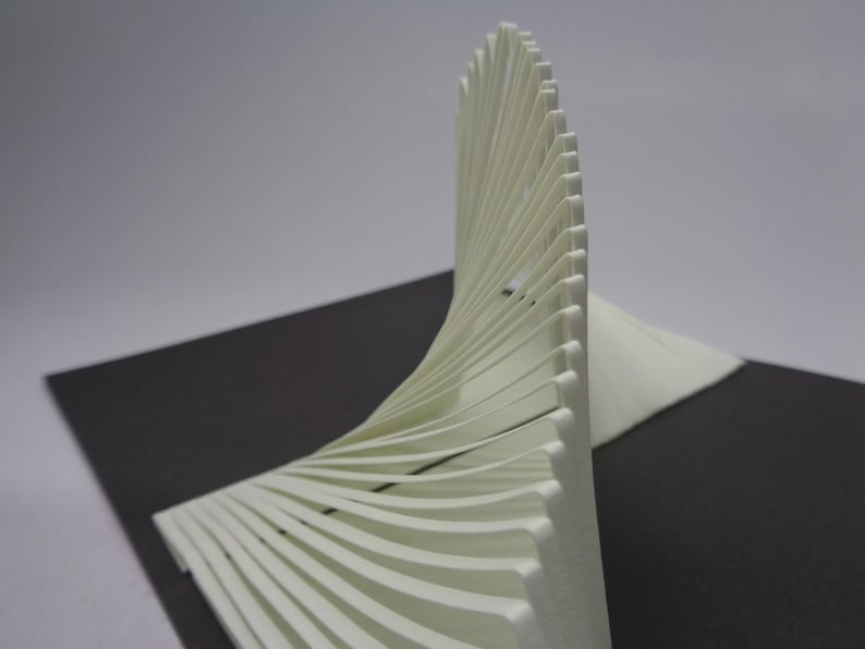 May include: A white, abstract sculpture with a curved, fan-like design. The sculpture is made of layered, thin, white material, creating a sense of depth and texture. It sits on a dark gray surface.