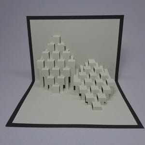 Puede incluir: Una tarjeta desplegable blanca con borde negro, que presenta dos formaciones cúbicas tridimensionales abstractas. Los cubos están dispuestos en un patrón en capas, creando un diseño geométrico. La tarjeta está abierta, mostrando los elementos desplegables.