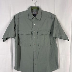 May include: A short-sleeved, sage green button-down shirt with two chest pockets. The shirt has a collared neckline and epaulets on the shoulders. It is hanging on a metal hanger against a white background.