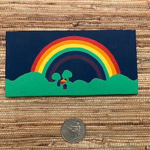 May include: A small, rectangular painting featuring a vibrant rainbow arching over a green landscape with two trees and small houses. The background is a deep blue, and the artwork is set against a textured, neutral background.