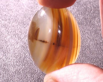 May include: An oval-shaped gemstone with a polished surface. The stone features bands of brown, tan, and cream colors. A dark line runs across the center of the stone. The gemstone is held between two fingers.
