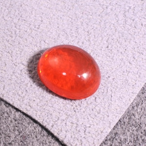 Puede incluir: Una piedra preciosa de forma ovalada y lisa con un tono naranja vibrante y translúcido. La piedra presenta una superficie pulida y reflectante que capta la luz, mostrando un color naranja rojizo profundo y uniforme en su forma redondeada.
