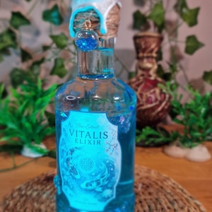 Può includere: Una bottiglia di vetro blu riempita di liquido blu, etichettata "VITALIS ELIXIR". La bottiglia è sigillata con una sostanza cerosa bianca e blu e spago. Una piccola sfera di vetro con glitter blu è appesa in cima. La bottiglia è su un tappetino intrecciato.