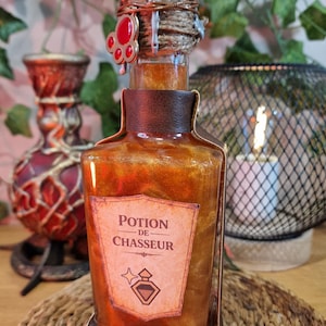 Peut inclure: Une bouteille de potion décorative avec un cadre en similicuir marron et un dessus argenté. La bouteille contient un liquide doré scintillant et porte une étiquette « Potion de Chasseur ». Une gemme rouge orne le dessus.