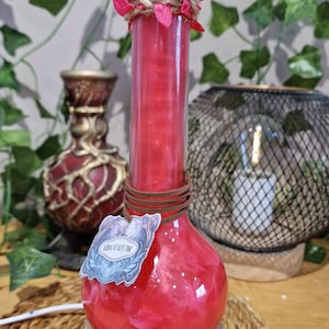 Può includere: Una bottiglia in vetro trasparente riempita con un liquido rosso scintillante, poggiata su una base rotonda in legno. Il collo è avvolto da spago e foglie finte rosa, con un cordino marrone che regge un'etichetta con scritto Vitality Tonic.