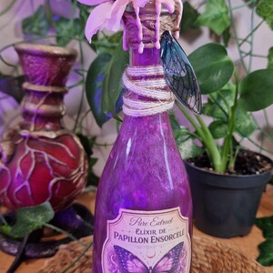 Op de afbeelding: Een paarse glazen fles met een bloem- en vlindervleugeldecoratie. De fles is gelabeld met "Pure Extract Élixir de Papillon Ensorcelé Mystical Tonic". De fles is omwikkeld met touw en bekroond met een roze bloem.