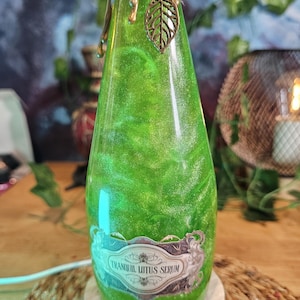 Può includere: Una bottiglia di vetro riempita con un liquido verde scintillante, con un'etichetta che riporta la scritta TRANQUIL LITUS SERUM. Il collo è avvolto da spago e sigillato con cera dorata, arricchito da un piccolo ciondolo a forma di foglia.