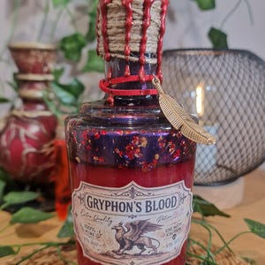 Peut inclure: Une bouteille en verre remplie de liquide rouge avec une étiquette indiquant GRYPHON'S BLOOD, Extra Quality, Potion No 39, 100% PURE, USE WITH CAUTION, ETS 1837. Le goulot est entouré de ficelle et de cire rouge, avec une breloque plume dorée.