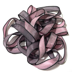 May include: A pile of sheer ribbon in shades of pink and gray, with black edging and white stitching. The ribbon is loosely arranged, creating a textured and layered effect. Ideal for crafts and gift wrapping.