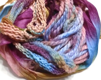 May include: A close-up of a ball of colorful yarn and ribbons. The yarn is a mix of pink, blue, purple, and tan, with a variety of textures. The ribbons are silk and satin in shades of blue, purple, and gold.