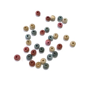 May include: A collection of small, round wooden beads in various muted colors, including pink, gray, beige, and green. The beads are scattered on a white surface, showcasing their natural texture and hole through the center.