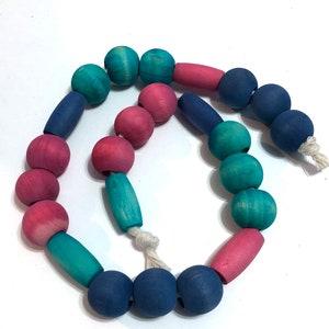 May include: A colorful wooden bead garland featuring a variety of shapes and colors. The beads are painted in shades of blue, pink, and teal, strung together with a natural fiber cord. The garland is a decorative item.