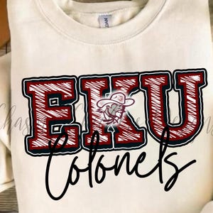 May include: Cream-colored sweatshirt featuring the text "EKU Colonels" in a bold, outlined design. The letters are red with a textured effect, and the logo includes a cowboy. The word "Colonels" is written in a cursive font.