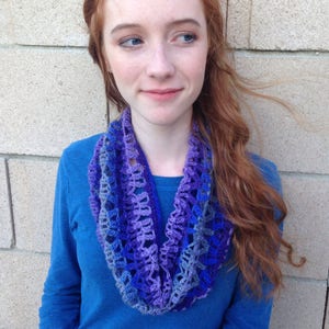 May include: A woman wearing a blue long-sleeved shirt with a purple, blue and grey crocheted infinity scarf.