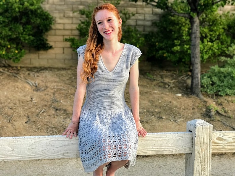 Puede incluir: Una mujer lleva un vestido de crochet gris claro con cuello en V y mangas cortas. El vestido tiene un patr&oacute;n de encaje de crochet en la falda.