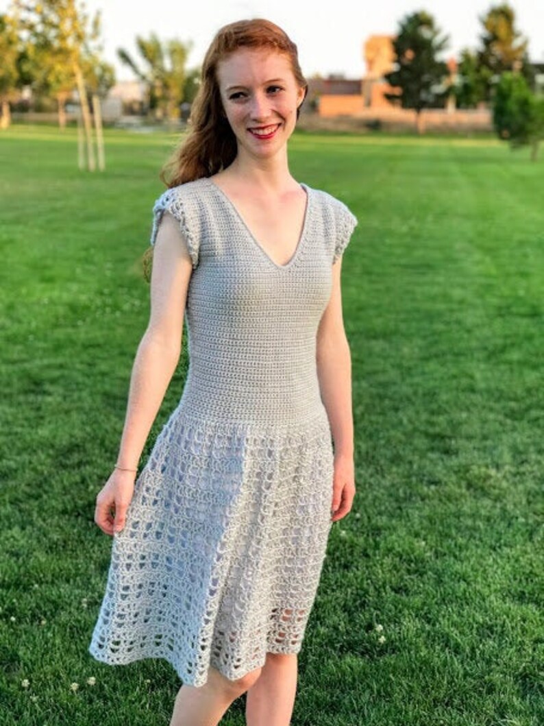 Puede incluir: Un vestido de crochet gris claro con cuello en V y mangas cortas. El vestido tiene un corpi&ntilde;o ajustado y una falda fluida.