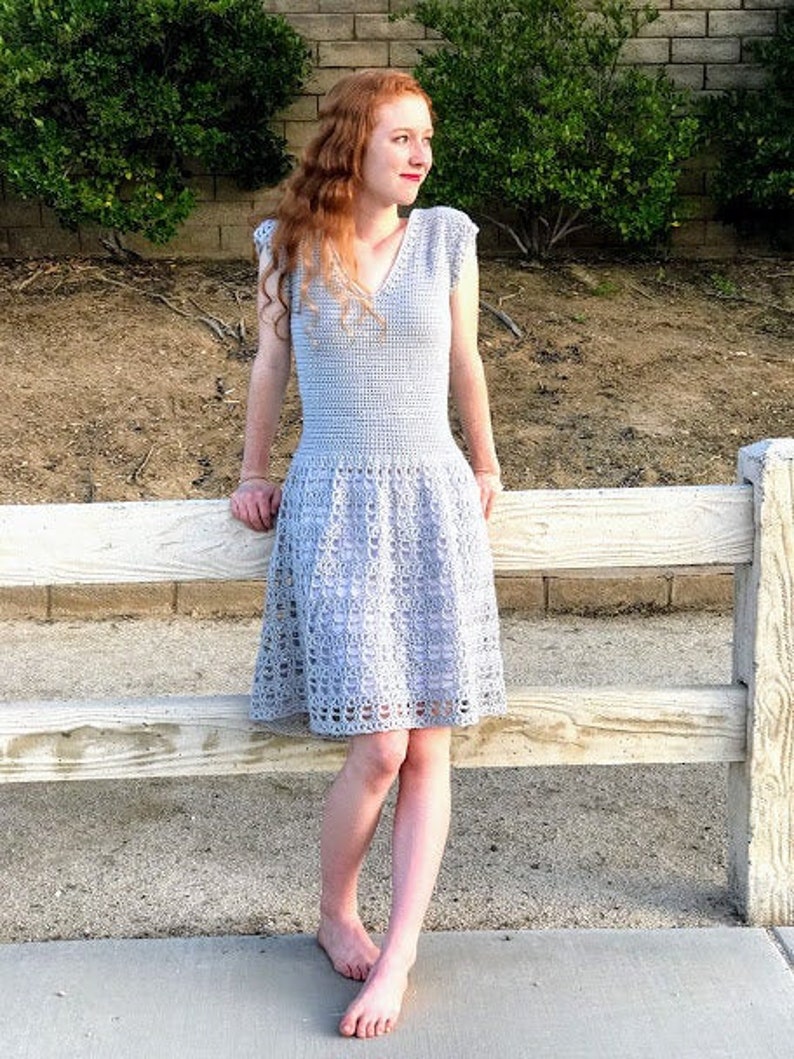 Puede incluir: Una mujer lleva un vestido de crochet gris claro con cuello en V y mangas cortas. El vestido tiene un corpi&ntilde;o ajustado y una falda fluida. Est&aacute; de pie frente a una valla de madera blanca.