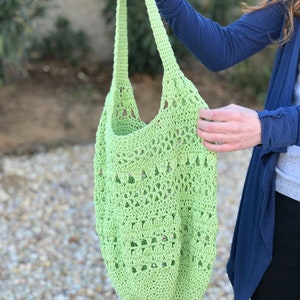 Puede incluir: Una bolsa de mano de crochet de color verde claro con un patr&oacute;n delicado. La bolsa tiene una sola correa y es lo suficientemente grande como para llevar comestibles u otros art&iacute;culos.