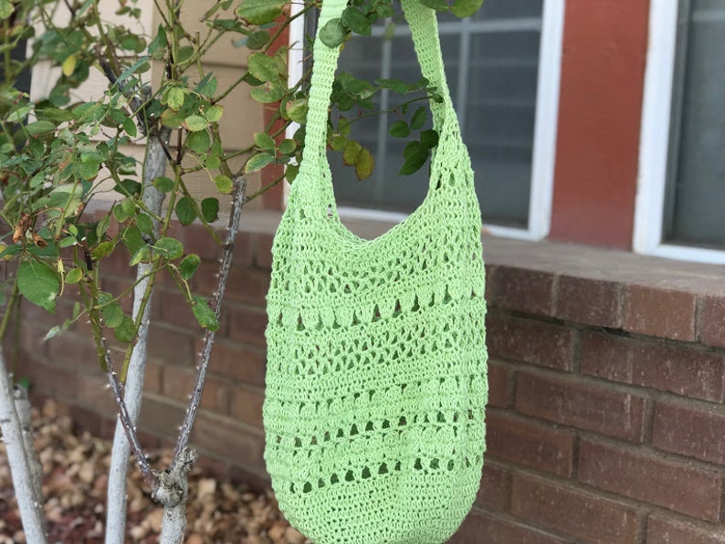 Puede incluir: Un bolso tote de crochet de color verde claro con una correa larga. El bolso tiene un delicado patr&oacute;n de calado y est&aacute; colgado de una rama de &aacute;rbol.