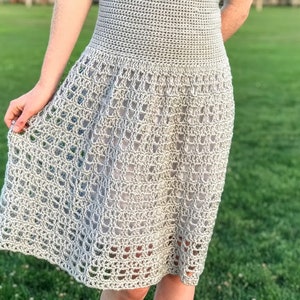 Puede incluir: Un vestido de crochet gris claro con un corpi&ntilde;o ajustado y una falda fluida. La falda tiene un delicado patr&oacute;n de calado.