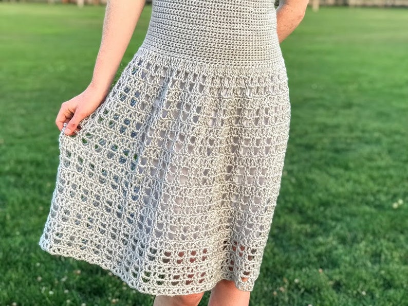 Puede incluir: Un vestido de crochet gris claro con un corpi&ntilde;o ajustado y una falda fluida. La falda tiene un delicado patr&oacute;n de calado.