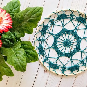 May include: A teal crocheted dreamcatcher with a wooden hoop, hanging against a white wood background.