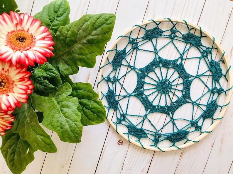 May include: A teal crocheted dreamcatcher with a wooden hoop, hanging against a white wood background.