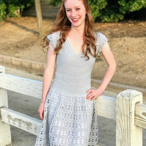 Puede incluir: Una mujer lleva un vestido de crochet gris con un dobladillo festoneado. El vestido tiene un escote en V y mangas cortas.