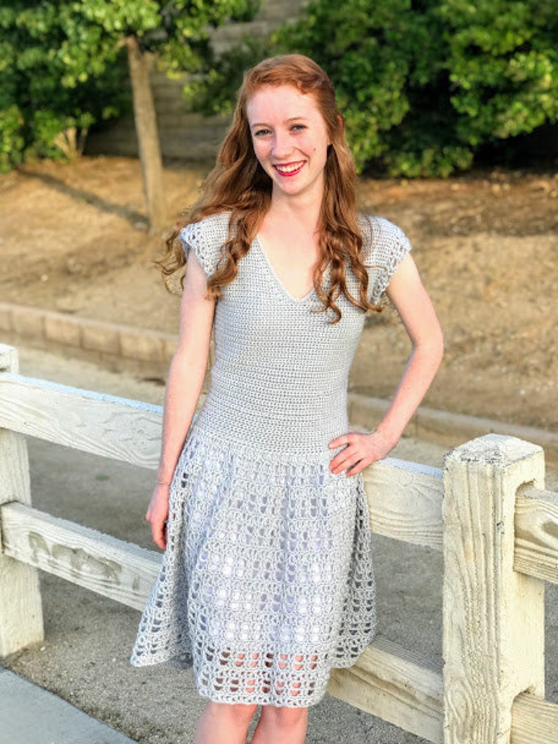 Puede incluir: Una mujer lleva un vestido de crochet gris con un dobladillo festoneado. El vestido tiene un escote en V y mangas cortas.