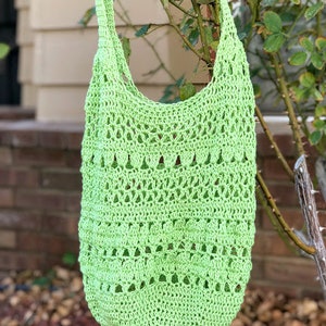 Puede incluir: Una bolsa de mano de crochet de color verde lima con una parte superior ancha y abierta y una correa larga. La bolsa est&aacute; colgada de una rama de un arbusto.