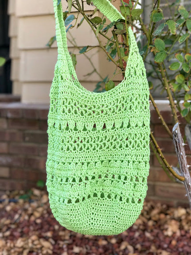 Puede incluir: Una bolsa de mano de crochet de color verde lima con una parte superior ancha y abierta y una correa larga. La bolsa est&aacute; colgada de una rama de un arbusto.