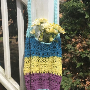 Puede incluir: Una bolsa de mano de crochet con rayas azules, amarillas y moradas. La bolsa est&aacute; colgada de un poste de madera blanco y tiene una correa azul claro. Hay flores blancas dentro de la bolsa.