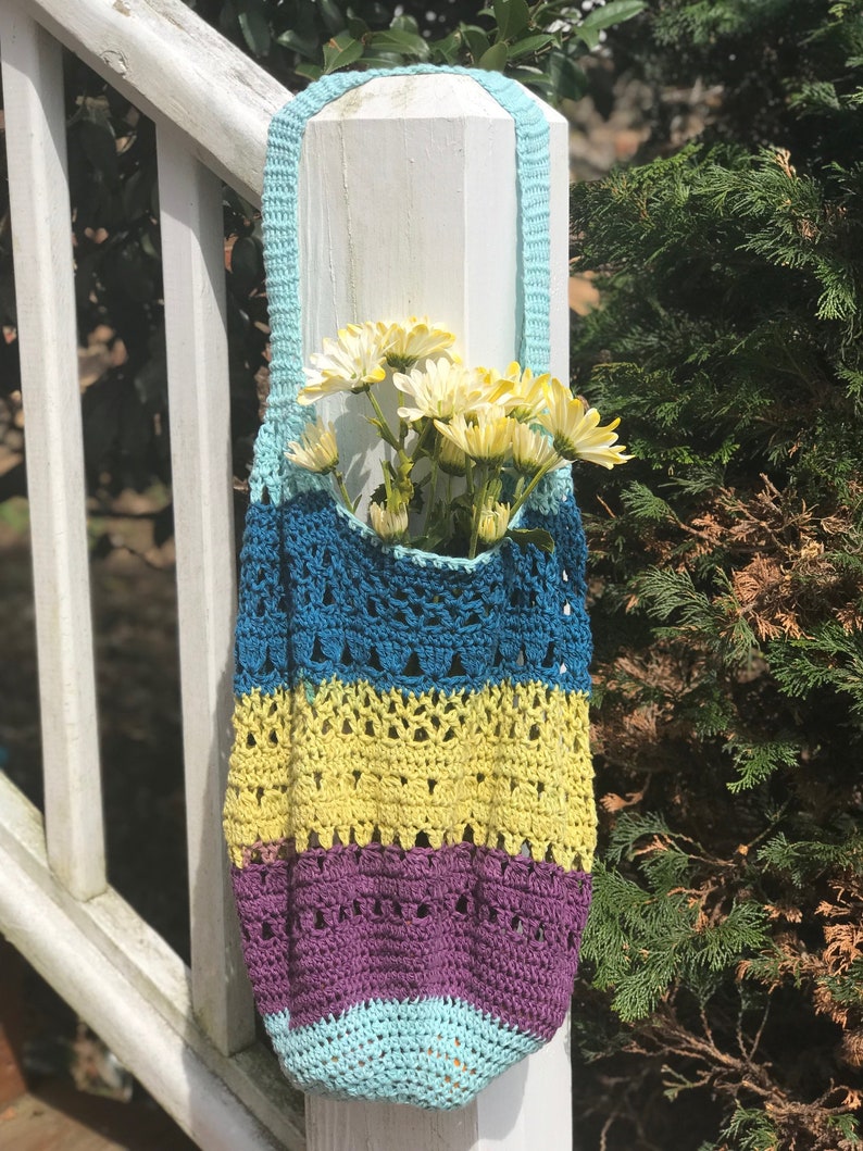 Puede incluir: Una bolsa de mano de crochet con rayas azules, amarillas y moradas. La bolsa est&aacute; colgada de un poste de madera blanco y tiene una correa azul claro. Hay flores blancas dentro de la bolsa.