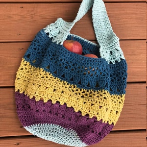 Puede incluir: Una bolsa de mano de crochet con un dise&ntilde;o a rayas azul, amarillo y morado. La bolsa tiene una sola correa larga y est&aacute; llena de manzanas rojas.