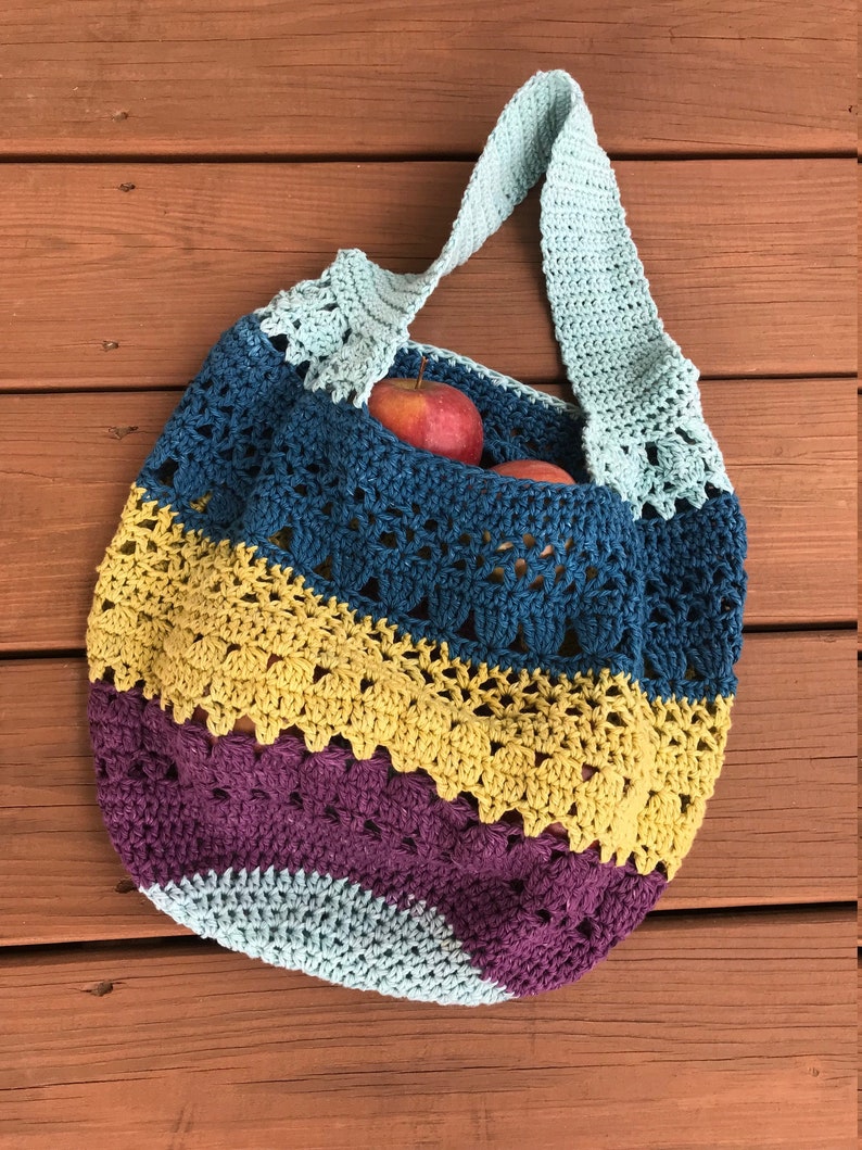 Puede incluir: Una bolsa de mano de crochet con un dise&ntilde;o a rayas azul, amarillo y morado. La bolsa tiene una sola correa larga y est&aacute; llena de manzanas rojas.