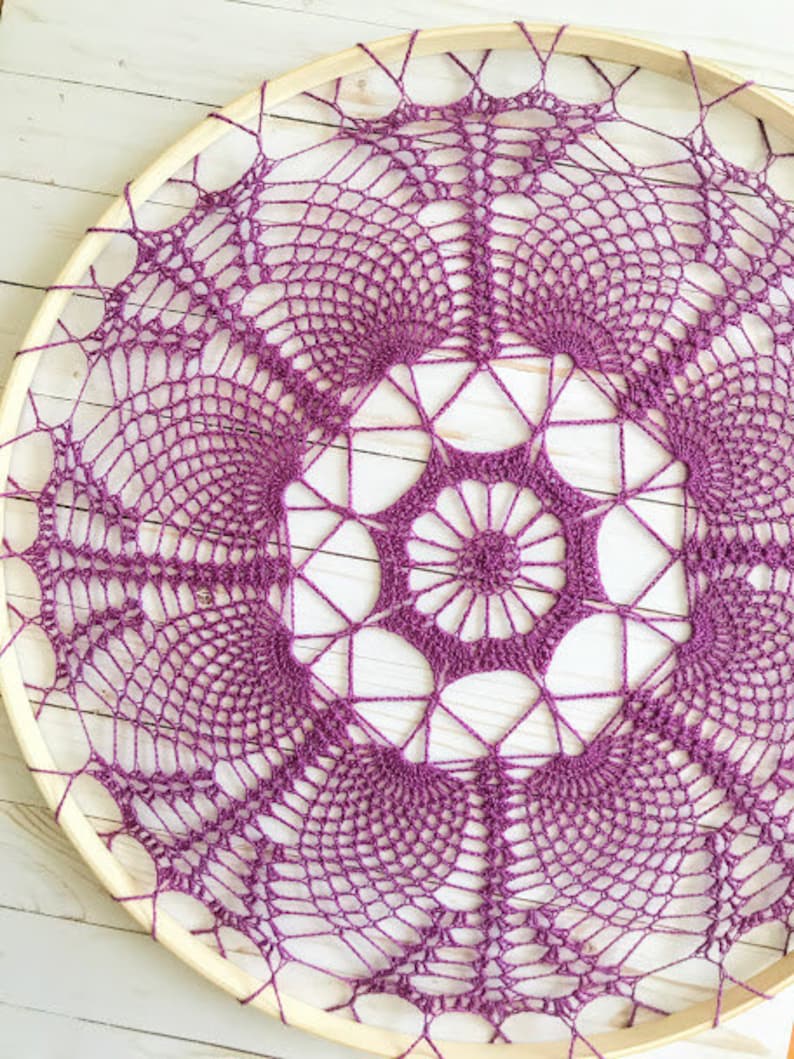 May include: A purple crochet doily is stretched over a wooden embroidery hoop. The doily has a delicate, intricate design with a central flower motif.