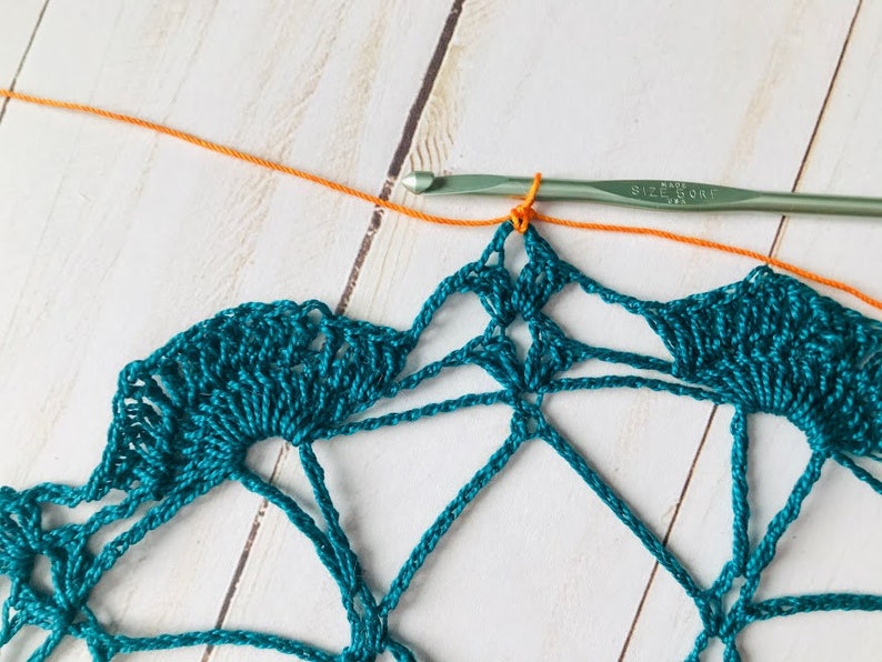 May include: A teal crochet doily with a single orange yarn strand attached to the hook. The hook is labeled "Size H/8 (5.0 mm)".