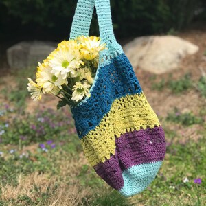 Puede incluir: Una bolsa de mano de crochet con un dise&ntilde;o a rayas azul, turquesa, amarillo y morado. La bolsa est&aacute; llena de un ramo de margaritas blancas.