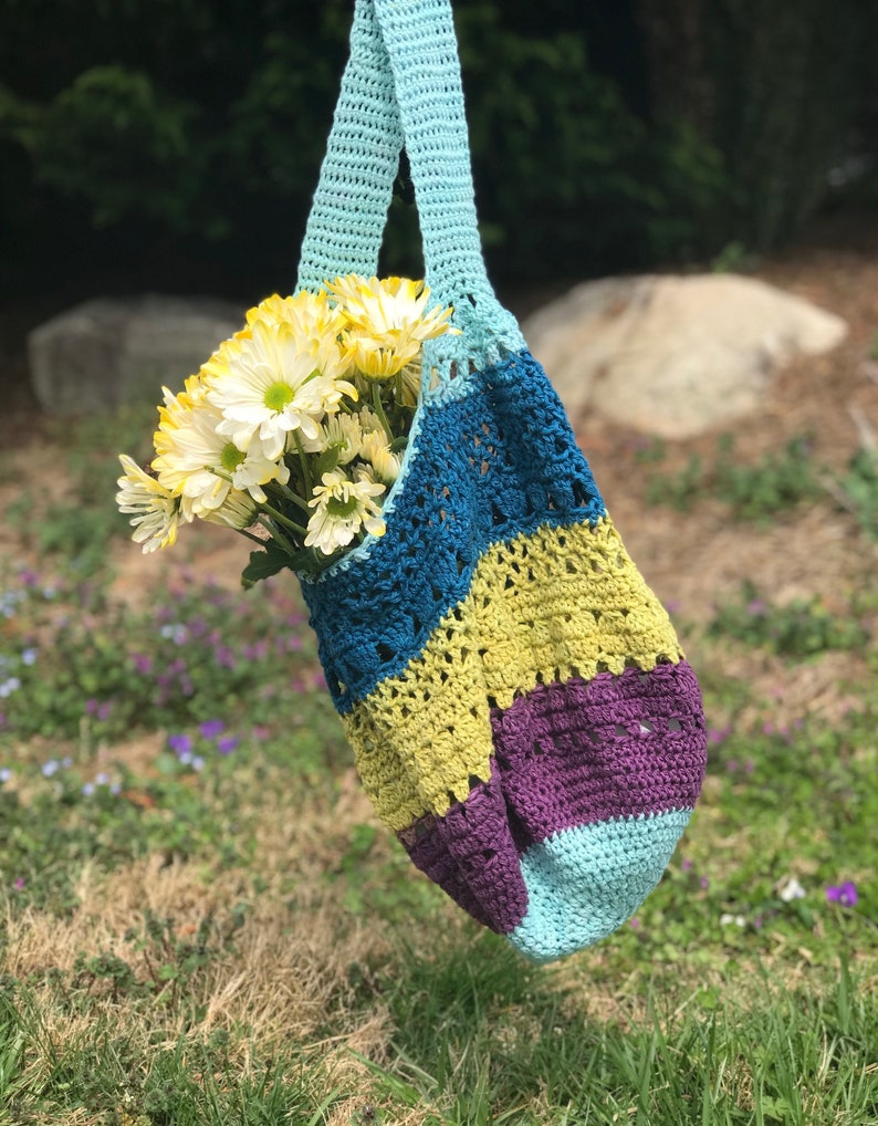Puede incluir: Una bolsa de mano de crochet con un dise&ntilde;o a rayas azul, turquesa, amarillo y morado. La bolsa est&aacute; llena de un ramo de margaritas blancas.