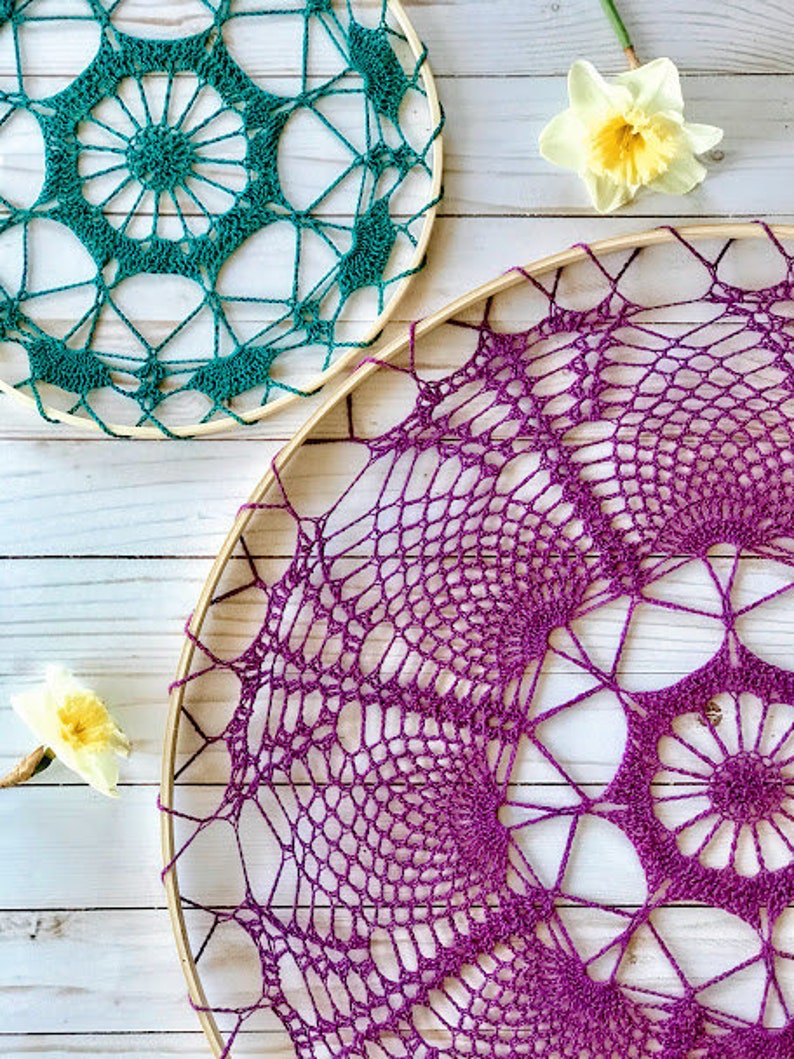 May include: Two crocheted dreamcatchers, one teal and one purple, hanging on a white wooden background.