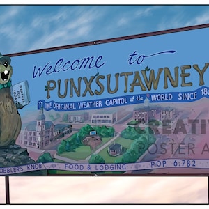 Puede incluir: Un letrero azul y blanco con el texto "Bienvenido a Punxsutawney" y una marmota de dibujos animados sosteniendo un libro y un paraguas. El letrero también incluye el texto "The Original Weather Capitol of the World Since 1887", "Gobbler's Knob", "Food & Lodging", y "Pop. 6,782".