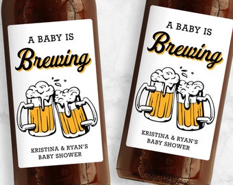 May include: Two brown beer bottles with white labels. The labels have a black and yellow illustration of two beer mugs with foam on top. The text on the labels reads "A Baby Is Brewing" and "Kristina & Ryan's Baby Shower".