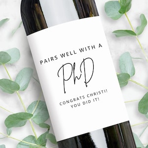 May include: A white wine bottle label with black text that reads "Pairs well with a PhD. Congrats Christi! You did it!"
