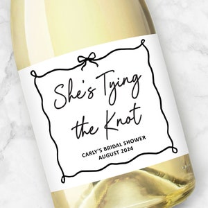 May include: White wine bottle with a black and white label that says "She's Tying the Knot" with a bow at the top. The label also says "Carly's Bridal Shower August 2024".