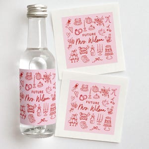 May include: A clear glass bottle with a silver cap and a pink label featuring red illustrations and the text "FUTURE Mrs. Wilson." Two white square coasters with matching pink and red designs are also present. Wedding theme.
