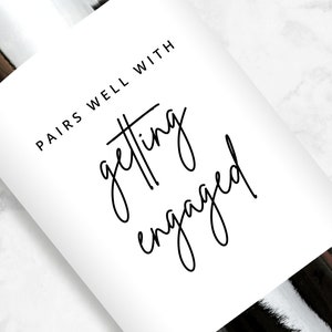 May include: White wine bottle label with black text that reads "Pairs well with getting engaged".