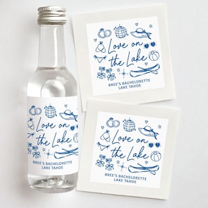 May include: A clear glass bottle with a silver cap and a white label featuring blue illustrations and text. The label reads "Love on the Lake" and "Bree's Bachelorette Lake Tahoe." Two square cards with the same design are also visible.