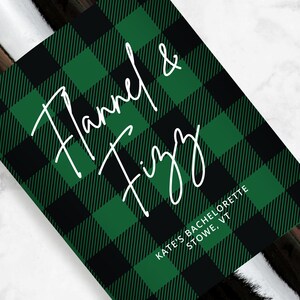 May include: Green and black buffalo plaid wine bottle label with white text that reads "Flannel & Fizzy" and "Kate's Bachelorette Stowe, VT".
