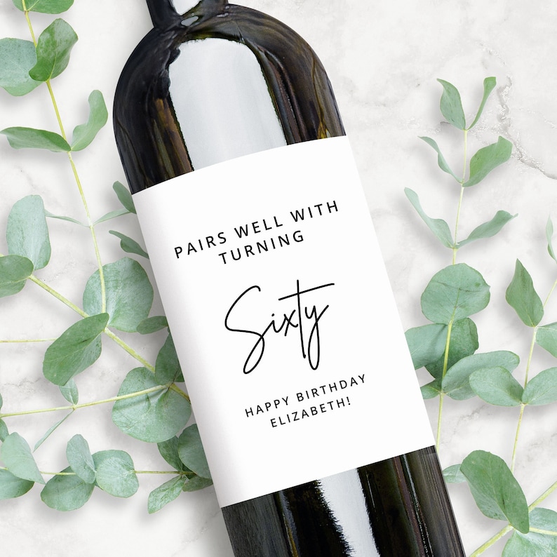 May include: A white wine bottle label with black text that reads "Pairs well with turning Sixty. Happy Birthday Elizabeth!"